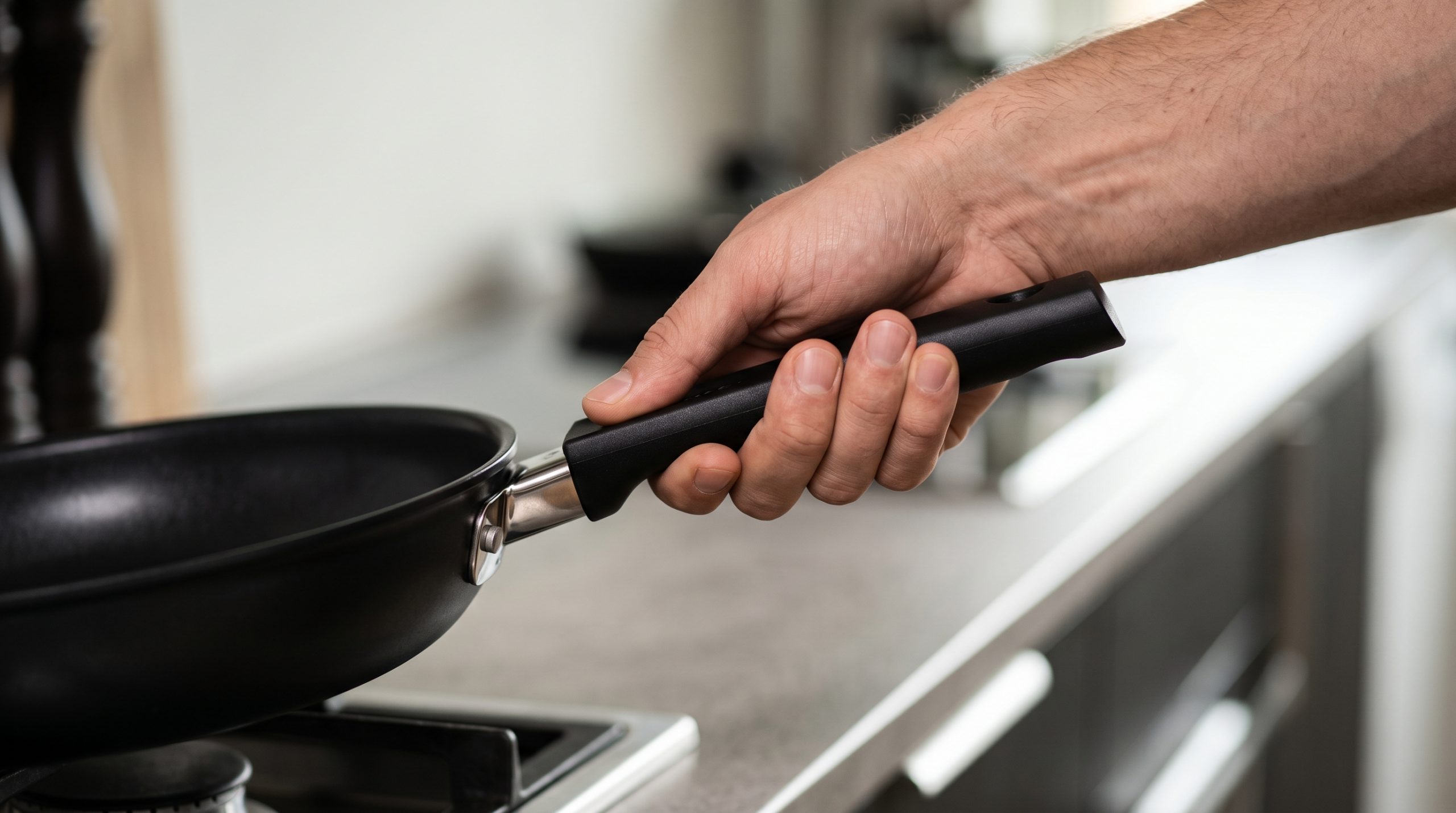 Närbild på en hand som håller ett stadigt och bekvämt grepp om det svarta, ergonomiska handtaget på en stekpanna. I den oskarpa bakgrunden syns ett modernt kök